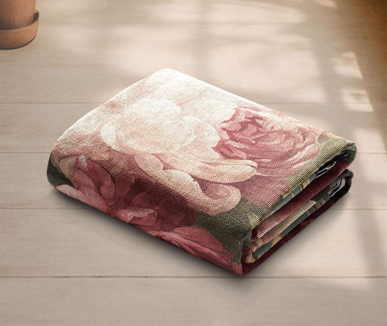 K&ouml;nnte beinhalten: Gefaltete Blumendecke mit einem gro&szlig;en Rosenmuster in Rosa, Creme und Gr&uuml;n. Die Decke ist ordentlich gefaltet und zeigt das detaillierte Design und die Textur des Stoffes. Die Decke liegt auf einer hellen Holzoberfl&auml;che.