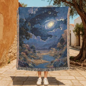 May include: A blue woven throw blanket featuring a celestial landscape with a galaxy, clouds, and trees. The blanket has a fringed edge and is held up by a person wearing white sneakers. The color palette includes blues, greens, and yellows.