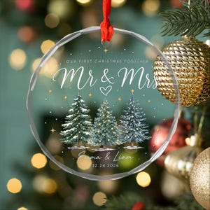 May include: A round, clear acrylic Christmas ornament featuring the text "OUR FIRST CHRISTMAS TOGETHER", "Mr & Mrs", and the names "Emma & Liam" with the date "12.24.2026". The design includes three illustrated pine trees and gold stars.