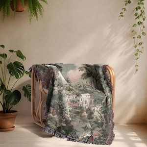 Pu&ograve; includere: Una coperta tessuta drappeggiata su una sedia in rattan. La coperta presenta una scena paesaggistica dettagliata nei toni del verde, rosa e bianco, con un bordo a frange. La sedia &egrave; in rattan marrone chiaro.