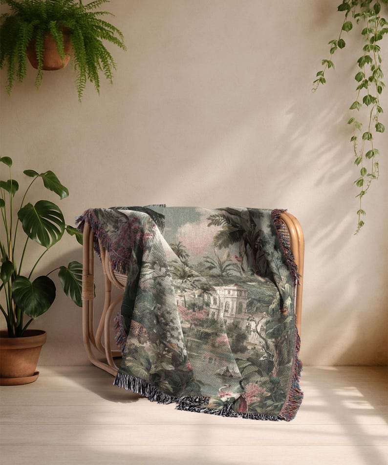 Pu&ograve; includere: Una coperta tessuta drappeggiata su una sedia in rattan. La coperta presenta una scena paesaggistica dettagliata nei toni del verde, rosa e bianco, con un bordo a frange. La sedia &egrave; in rattan marrone chiaro.
