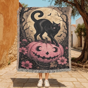 Könnte beinhalten: Eine gewebte Wohndecke mit einem schwarzen Katzenmotiv, die auf einem großen rosa Kürbis steht. Der Hintergrund zeigt einen Vollmond, kahle Bäume, Spinnenweben und Fledermäuse. Das Design enthält rosa Blumen am unteren Rand und Fransen.