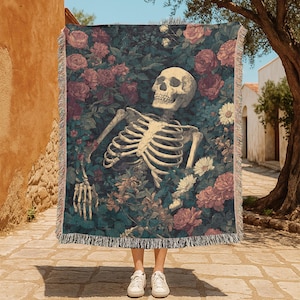 Könnte beinhalten: Eine gewebte Decke mit einem Skelett-Design, umgeben von Rosen und Blumen in Teal-, Burgunder- und Cremetönen. Die Decke hat einen Fransenrand. Die Person, die die Decke hält, trägt weiße Turnschuhe. Misst etwa 150 cm x 200 cm.