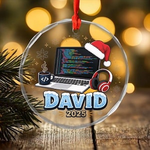 May include: A circular clear acrylic ornament featuring a laptop displaying colourful code, a coffee mug with a code symbol, headphones, and a red Santa hat. The name DAVID and the year 2025 are printed in blue with a white snowy effect.
