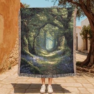 Op de afbeelding: Een geweven sprei met een gedetailleerde schildering van een bospad. Het kunstwerk toont een zonnig pad dat door weelderige groene bomen en blauwe bloemen slingert. De sprei heeft een gefranjerde rand.