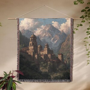 May include: Woven wall tapestry showing a mountain landscape with a medieval village. The tapestry features a detailed illustration of a stone village with domed towers and a cross on top. The colour palette includes shades of brown, green, and blue.