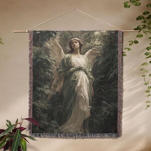 May include: A tapestry wall hanging featuring a classical painting of an angel with large wings, dressed in flowing robes, set against a backdrop of lush greenery. The artwork is framed with a fringed border and suspended from a wooden rod.