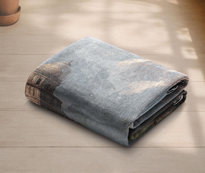 May include: Folded rectangular tapestry with a detailed architectural design, including a domed structure and a cloudy sky. The tapestry is mainly blue and brown, with visible folds and a textured appearance.