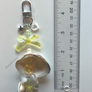 May include: A seashell keyring with a silver clasp, featuring a yellow bow, star charm, and assorted beads. The keyring includes a small seashell and is approximately 7.6 cm in length.