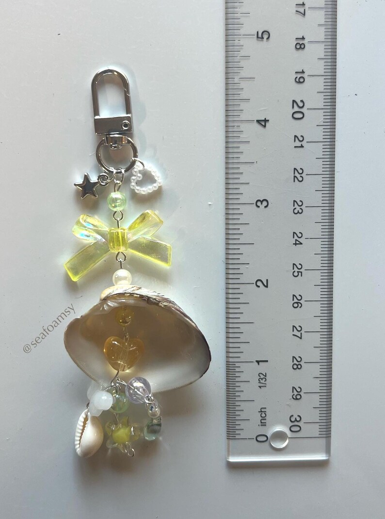 May include: A seashell keyring with a silver clasp, featuring a yellow bow, star charm, and assorted beads. The keyring includes a small seashell and is approximately 7.6 cm in length.