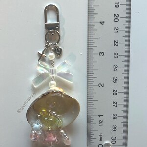 May include: A handmade keychain with a seashell, iridescent bow, and pastel-colored beads. The keychain has a silver-tone clasp, a star charm, and a heart charm. The ruler next to the keychain measures up to 76 cm.