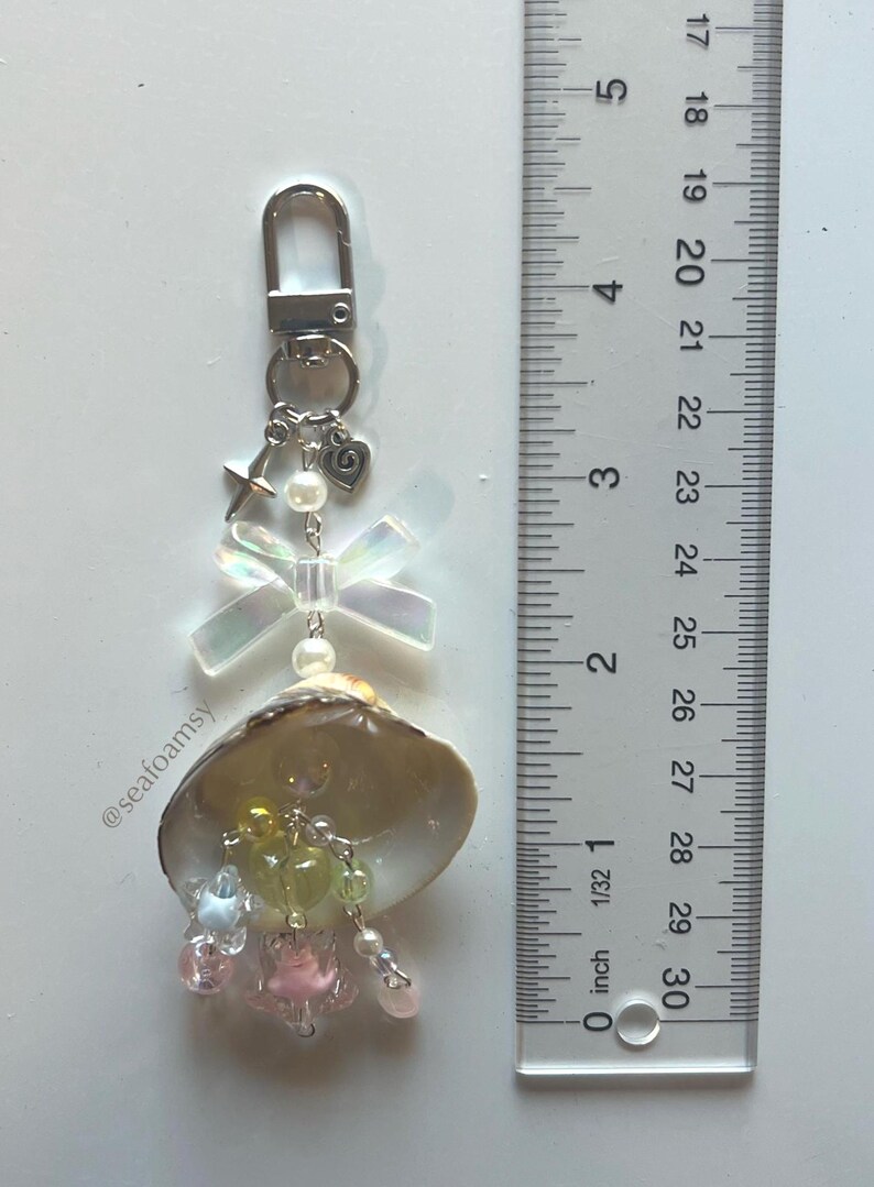 May include: A handmade keychain with a seashell, iridescent bow, and pastel-colored beads. The keychain has a silver-tone clasp, a star charm, and a heart charm. The ruler next to the keychain measures up to 76 cm.