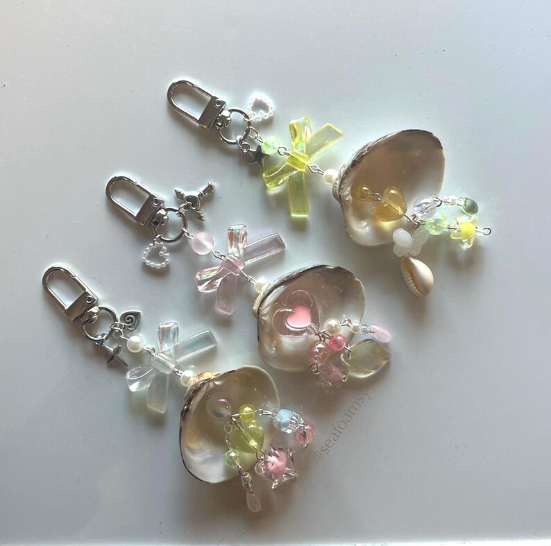 May include: Keyrings with seashell pendants, adorned with colourful beads and charms. Each keyring has a silver clasp and decorative elements such as crosses, hearts, and stars. The colour palette includes pink, yellow, and clear beads.