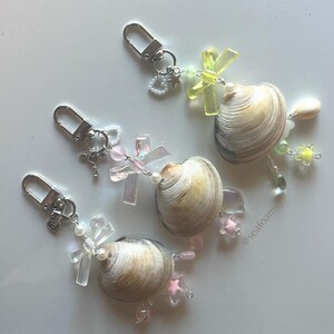 May include: Three seashell keyrings with silver clasps. Each keyring features a natural seashell, decorated with iridescent cross charms, pink star charms, and clear and pastel-coloured beads. The keyrings are arranged on a white surface.