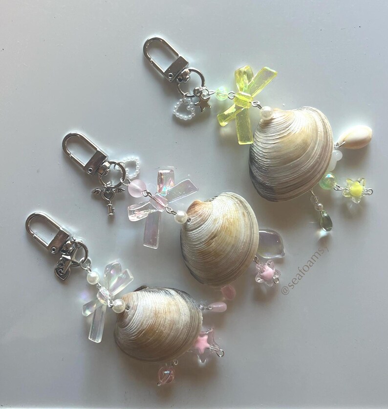 May include: Three seashell keyrings with silver clasps. Each keyring features a natural seashell, decorated with iridescent cross charms, pink star charms, and clear and pastel-coloured beads. The keyrings are arranged on a white surface.