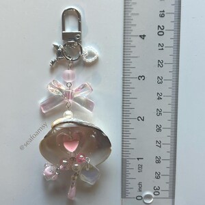 May include: A decorative keychain featuring a seashell with a pink heart, iridescent bow, and dangling pink beads. The keychain includes a silver clasp and a small key charm. The seashell is approximately 2.5 cm in length.
