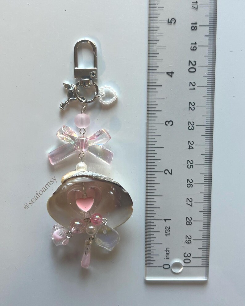 May include: A decorative keychain featuring a seashell with a pink heart, iridescent bow, and dangling pink beads. The keychain includes a silver clasp and a small key charm. The seashell is approximately 2.5 cm in length.