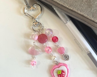 May include: A silver-coloured keyring with a heart-shaped clasp, featuring pink and red beads, a heart charm, and a strawberry pendant. The keyring is attached to a grey pencil case. The text "@seafoamsy" is visible.