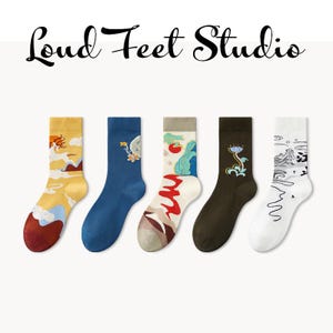 May include: Five pairs of colorful socks with unique designs, including illustrations of landscapes, flowers, and abstract patterns. The socks are in shades of yellow, blue, brown, and white, with the text "Loud Feet Studio" above.