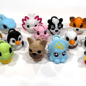 Op de afbeelding: Een verzameling kleurrijke sleutelhangers in de vorm van dieren, waaronder een konijn, een kikker, een haai, een varken, een vos, een hond, een pinguïn en een eend. Elke sleutelhanger heeft een kleine lus aan de bovenkant.