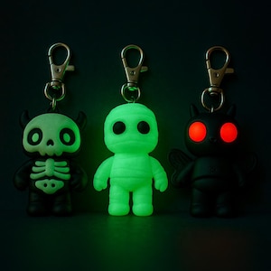 May include: Three Halloween-themed keychains. One is a black and white skeleton, the second is a green mummy, and the third is a black devil with glowing red eyes. Each keychain has a silver clasp.