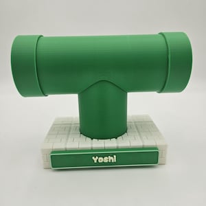 May include: A green T-shaped pipe display stand mounted on a white brick-patterned base. The front of the base features a green rectangular plaque with the word Yoshi printed in white letters. The structure has a smooth, matte finish throughout.
