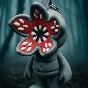 以下が含まれることがあります： 赤と白のアクセントと鋭い歯を持つ、花びらの形をした赤い頭を持つ灰色のデモゴルゴンフィギュア。フィギュアの頭にはキーリングが付いており、森の中に立っています。デモゴルゴンは人気のテレビ番組のキャラクターです。