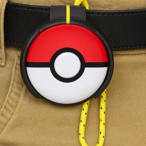 Op de afbeelding: Een rond, rood en wit accessoire met een zwarte rand, dat lijkt op een Pokébal, bevestigd aan een zwarte riem. Een geel koord met zwarte stippen hangt aan het accessoire. Het accessoire is bevestigd aan een kaki broek.