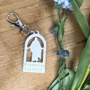 May include: A white acrylic keychain featuring an arched cutout silhouette of a figure with arms outstretched. Below the cutout, the text "Rejoice in CHRIST" is printed in white. The keychain is attached to a silver-toned metal lobster clasp.