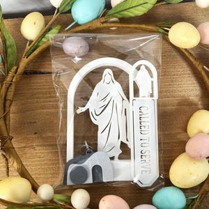May include: A white religious-themed keychain and bookmark set, packaged in clear plastic, featuring a figure and the text "CALLED TO SERVE." The set is displayed on a decorative wreath with pastel-colored eggs and white berries.