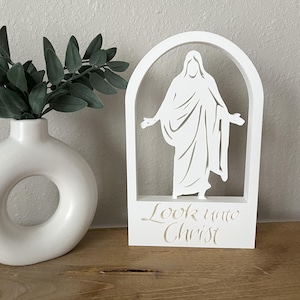 May include: A white wooden cutout of a religious figure within an arched frame. The figure has outstretched arms. The words "Look unto Christ" are cut out at the base. A white vase with green leaves is to the left.
