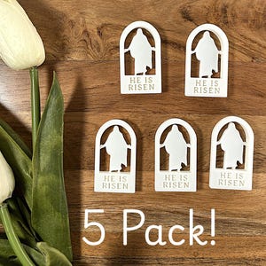 May include: A pack of five white, arched decorations with the text "HE IS RISEN" and a silhouette of a figure. The decorations are arranged on a wooden surface. The words "5 Pack!" are written below the decorations.