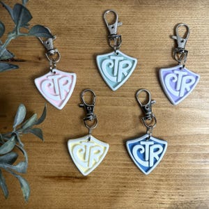 May include: Five shield-shaped keychains in pastel colors: pink, light blue, purple, yellow, and blue. Each keychain has a silver clasp and features the letters "CTR" in a contrasting color. The keychains are on a wooden surface.