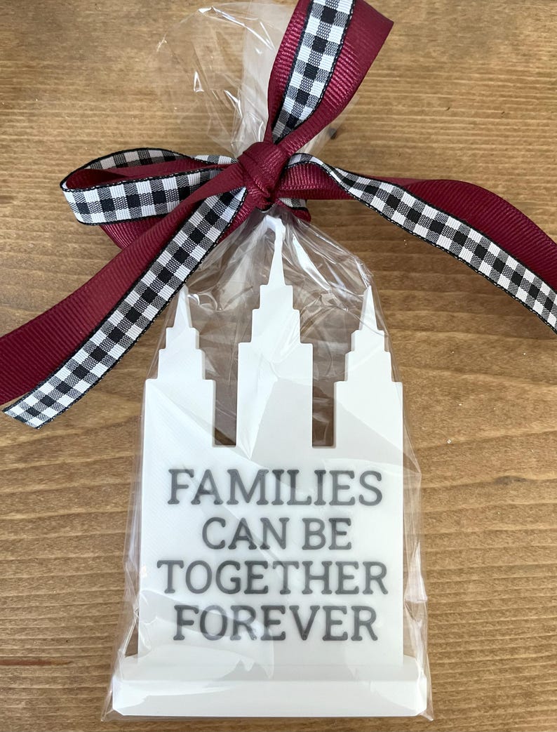 May include: A white, architectural-shaped ornament with the text "FAMILIES CAN BE TOGETHER FOREVER" in black, packaged in clear cellophane. The package is tied with a burgundy ribbon with a black and white checkered edge.