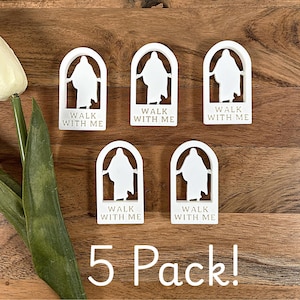 May include: Five white arched plaques with a silhouette of a figure inside. Each plaque has the words "WALK WITH ME" at the bottom. The text "5 Pack!" is below the plaques. The plaques are on a wooden surface.