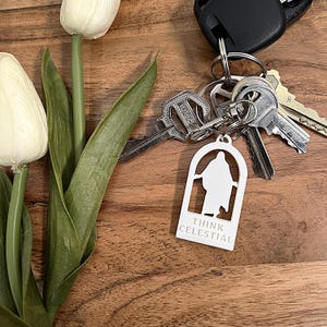 May include: White keychain with a cutout figure and the words "THINK CELESTIAL" below it. The keychain is attached to a set of keys. The background includes white tulips and a wooden surface.