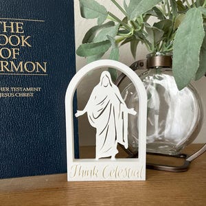 May include: A white, laser-cut decorative piece featuring a figure within an arched frame. The text "Think Celestial" is at the base. A dark blue book and a glass vase with greenery are in the background.