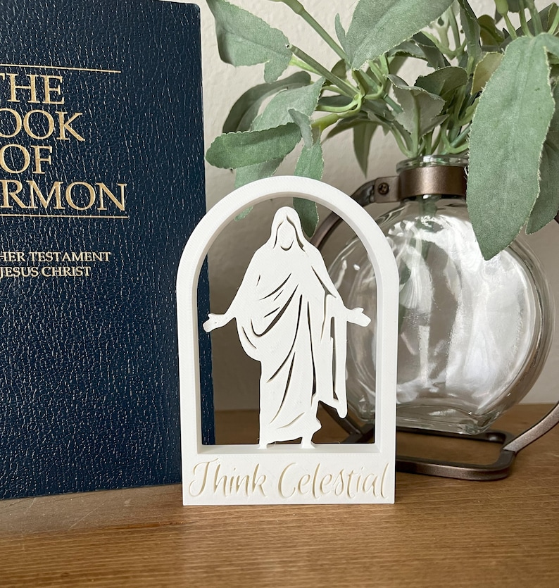 May include: A white, laser-cut decorative piece featuring a figure within an arched frame. The text "Think Celestial" is at the base. A dark blue book and a glass vase with greenery are in the background.