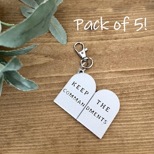 May include: A white heart-shaped keychain with the text "KEEP THE COMMANDMENTS" in black lettering. The keychain has a silver clasp and a speckled texture. The words "Pack of 5!" are written in white script.