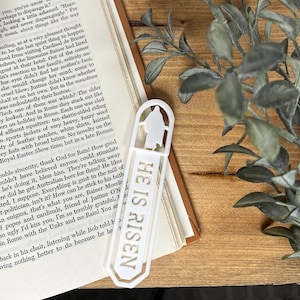 May include: A white acrylic bookmark with the text "HE IS RISEN" in a vertical orientation. The bookmark features a silhouette of a figure at the top and is placed on a wooden surface next to an open book and greenery.