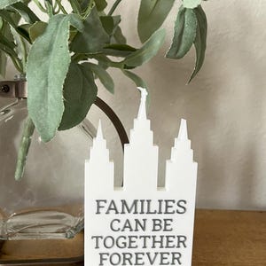 May include: A white decorative sign in the shape of a building with the text "FAMILIES CAN BE TOGETHER FOREVER". The sign is on a wooden surface, with greenery and a glass vase in the background.