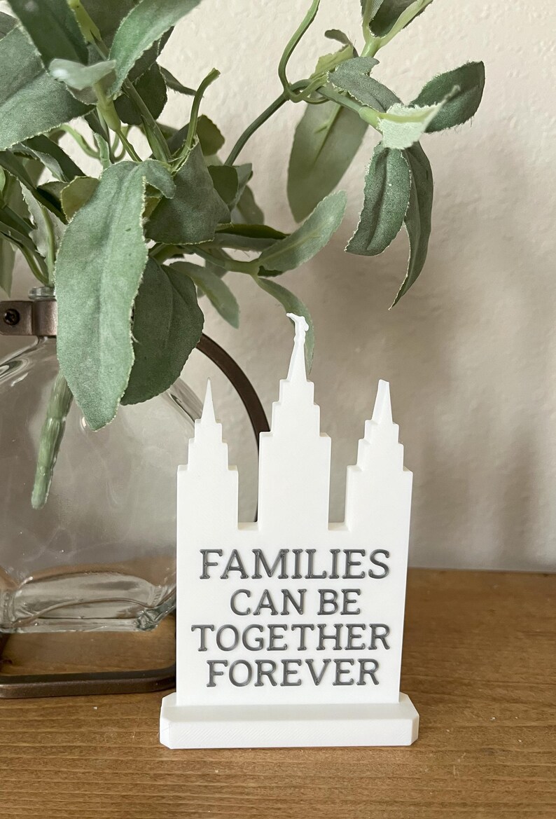May include: A white decorative sign in the shape of a building with the text "FAMILIES CAN BE TOGETHER FOREVER". The sign is on a wooden surface, with greenery and a glass vase in the background.