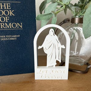 May include: A white, layered religious figurine depicting a figure within an arched frame, with the words "Let God Prevail" at the base. A dark blue book with gold lettering, titled "The Book of Mormon," is visible in the background.