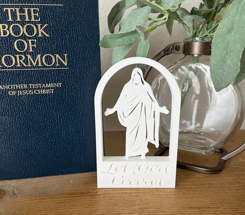 May include: A white, layered religious figurine depicting a figure within an arched frame, with the words "Let God Prevail" at the base. A dark blue book with gold lettering, titled "The Book of Mormon," is visible in the background.