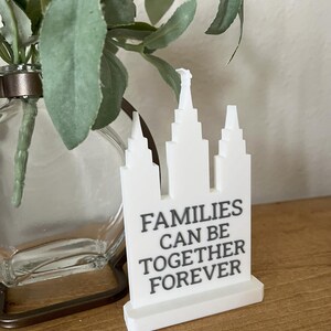 May include: A white decorative piece with a building design and the text "FAMILIES CAN BE TOGETHER FOREVER" in gray. The piece is on a wooden surface, with a glass vase and greenery in the background.