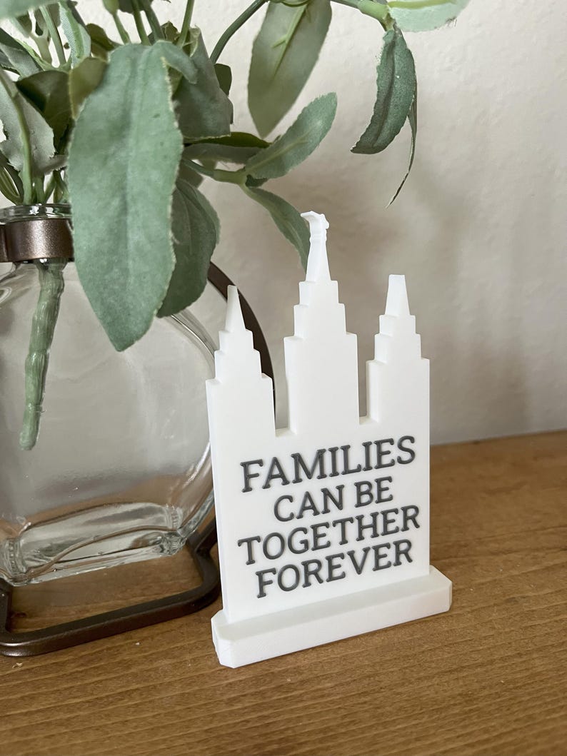 May include: A white decorative piece with a building design and the text "FAMILIES CAN BE TOGETHER FOREVER" in gray. The piece is on a wooden surface, with a glass vase and greenery in the background.