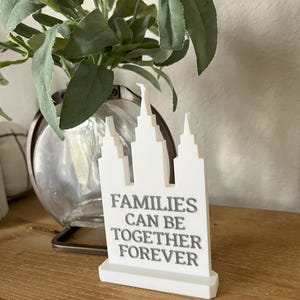 May include: A white decorative sign with a stylized building design and the text "FAMILIES CAN BE TOGETHER FOREVER" in gray. The sign is on a wooden surface, with a glass vase and greenery in the background.