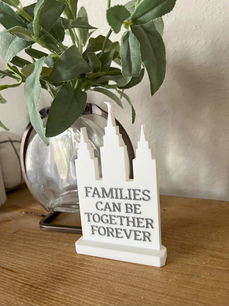 May include: A white decorative sign with a stylized building design and the text "FAMILIES CAN BE TOGETHER FOREVER" in gray. The sign is on a wooden surface, with a glass vase and greenery in the background.