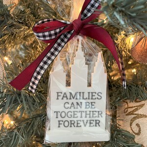 May include: A white wooden ornament in a clear cellophane bag, tied with a burgundy and black and white checkered ribbon. The ornament is shaped like a building with the text "FAMILIES CAN BE TOGETHER FOREVER" in gray.