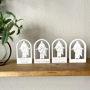 May include: Four white, arched figurines with a religious theme. Each features a figure with outstretched arms and text: "Look unto Christ", "Think Celestial", and "Let God Prevail". They are displayed on a wooden shelf with a plant in the background.